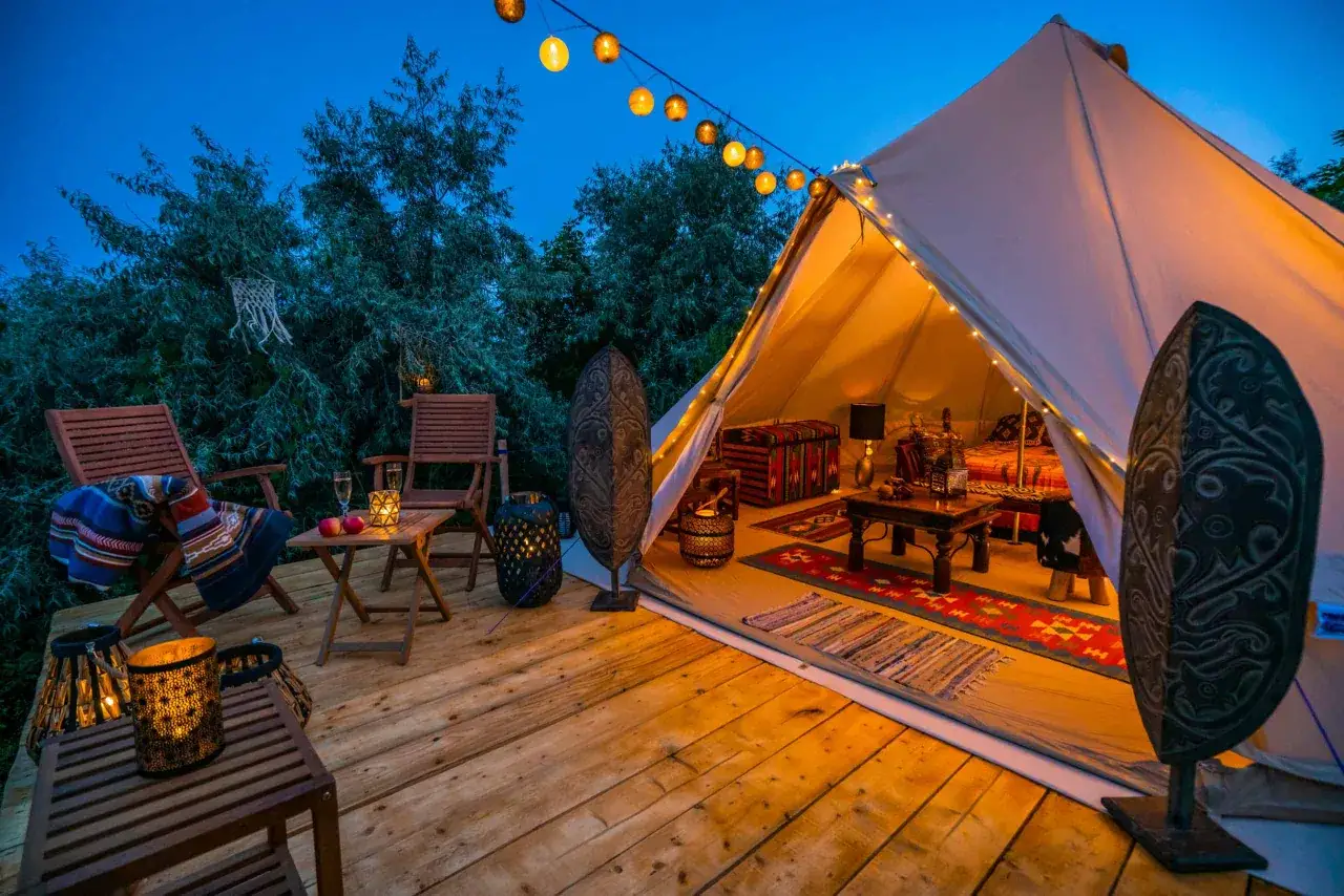 Experience glamping: a cozy, illuminated bell tent with a wooden deck, chairs, and decorative lanterns, set against a twilight sky and lush trees.