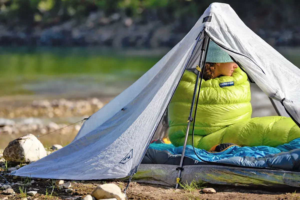 A person in a bright green sleeping bag sits in a tent by a river, contemplating the merits of a quilt vs. sleeping bag for their next adventure.