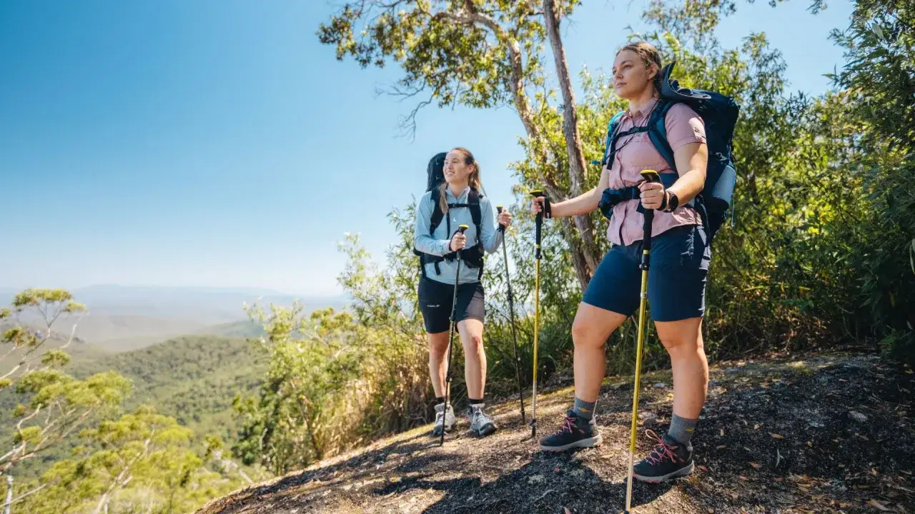 Two women hike on a sunny day, showcasing what to wear hiking in summer: shorts, light shirts, and sturdy boots.