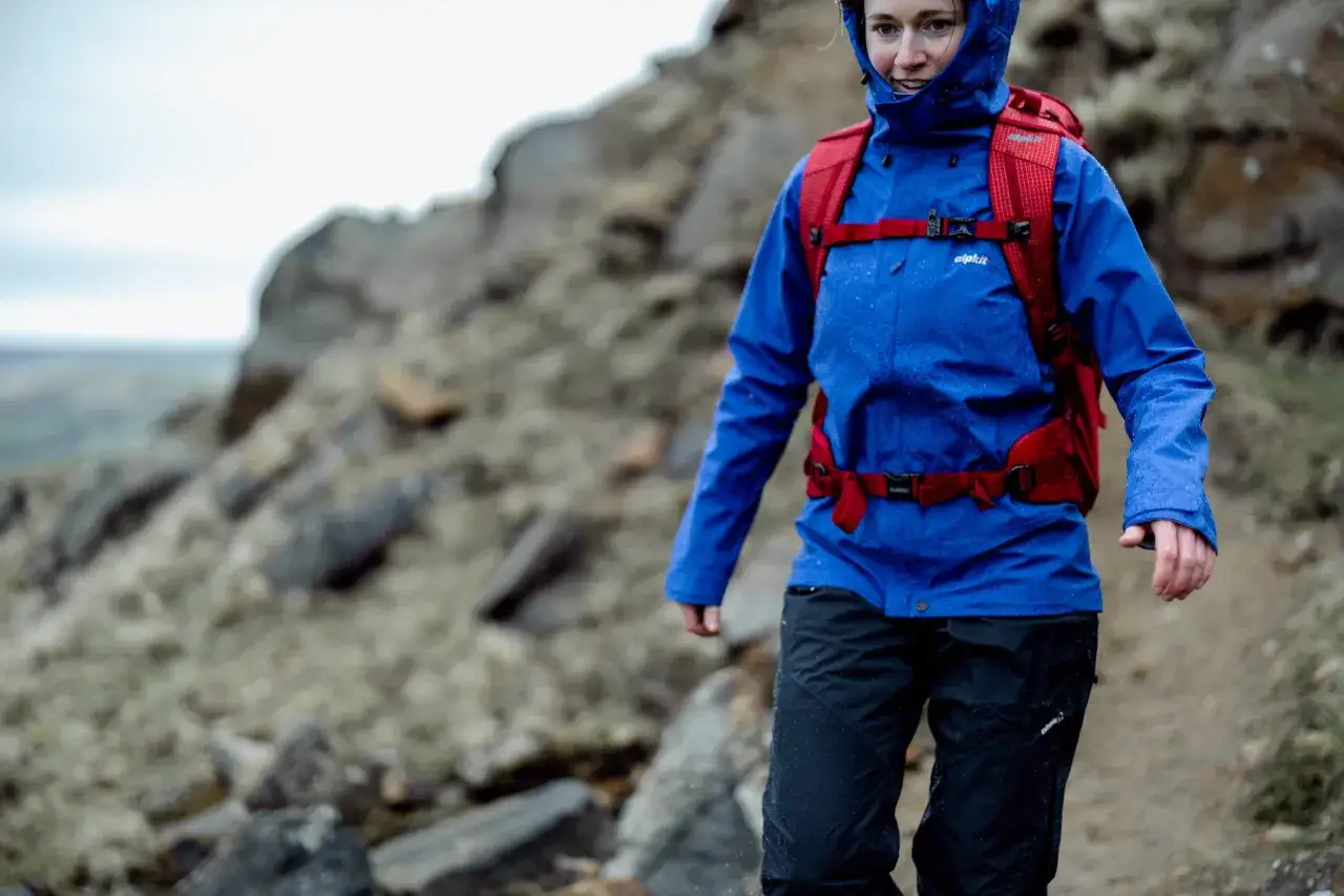 A woman in blue waterproof backpacking clothes and a red backpack walks on a rocky trail in the rain.