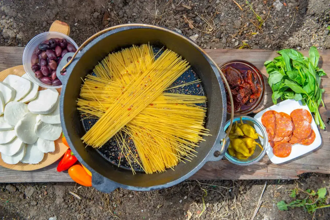 Gathering ingredients for delicious dutch oven campfire recipes: spaghetti, mozzarella, olives, sun-dried tomatoes, pepperoni, peppers, and basil.