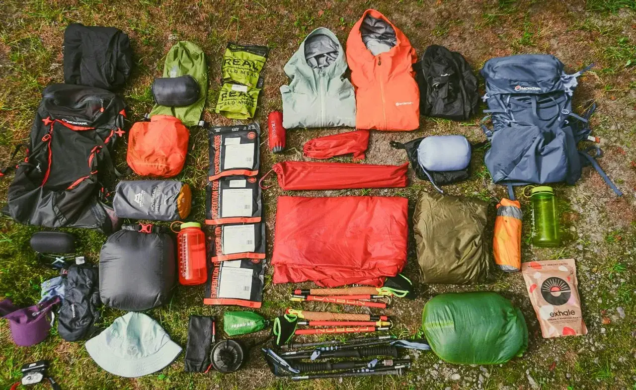 A comprehensive fastpacking gear list laid out on grass, featuring backpacks, sleeping bags, tents, jackets, food, and trekking poles.