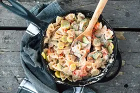 A hearty camping pasta dish with farfalle, zucchini, and tomatoes in a creamy sauce, served in a cast-iron skillet.