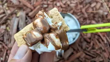 A hand holds a graham cracker topped with melted marshmallows and chocolate, ready to be assembled into a s'more. A pan of marshmallows is visible in the background.