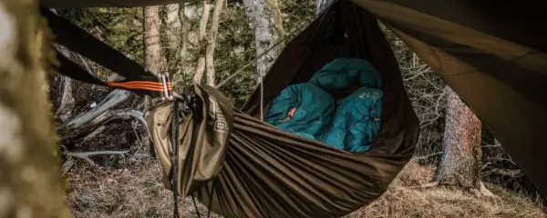 Cozy hammock camping setup in a forest. Two teal sleeping bags are nestled inside a brown hammock, ready for a night under the stars.