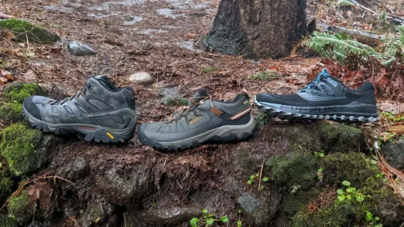 Three pairs of shoes on a wet forest floor: hiking boots, trail running shoes, and another pair of trail running shoes.