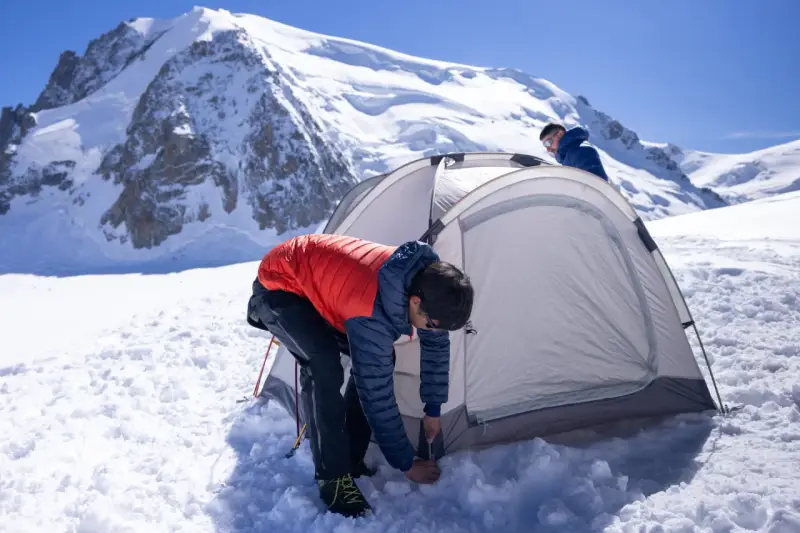 Two people set up a tent for winter camping amidst a snowy mountain landscape under a clear blue sky.