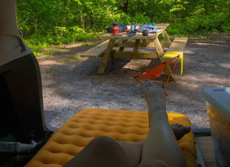 A person's legs rest on an orange sleeping pad inside a car. A picnic table with camping gear sits in a wooded area, ready for a road trip checklist.